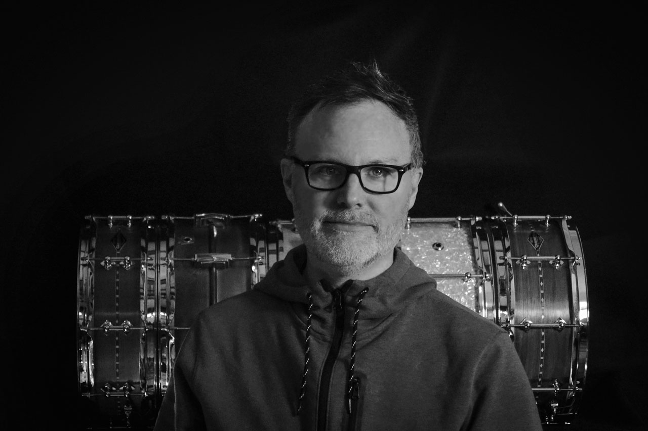 black and white portrait of ray in front of a shelf of a few snare drums